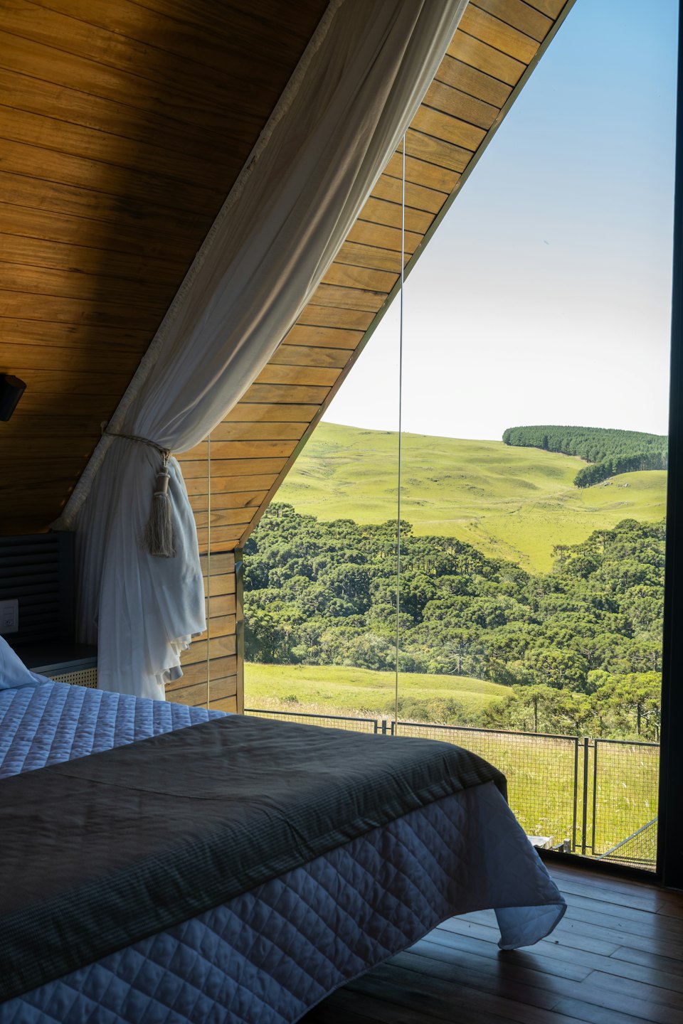 Bedroom with large triangular window overlooking green hills and forest.